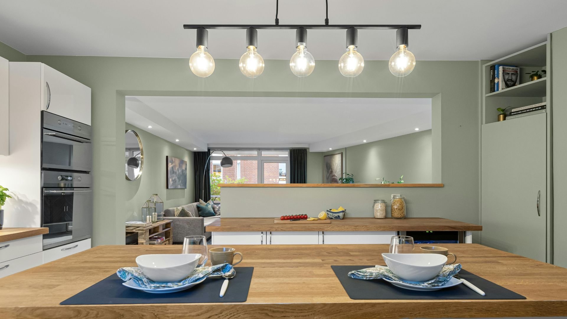 Modern kitchen with sage green walls, wooden table, and hanging light bulbs